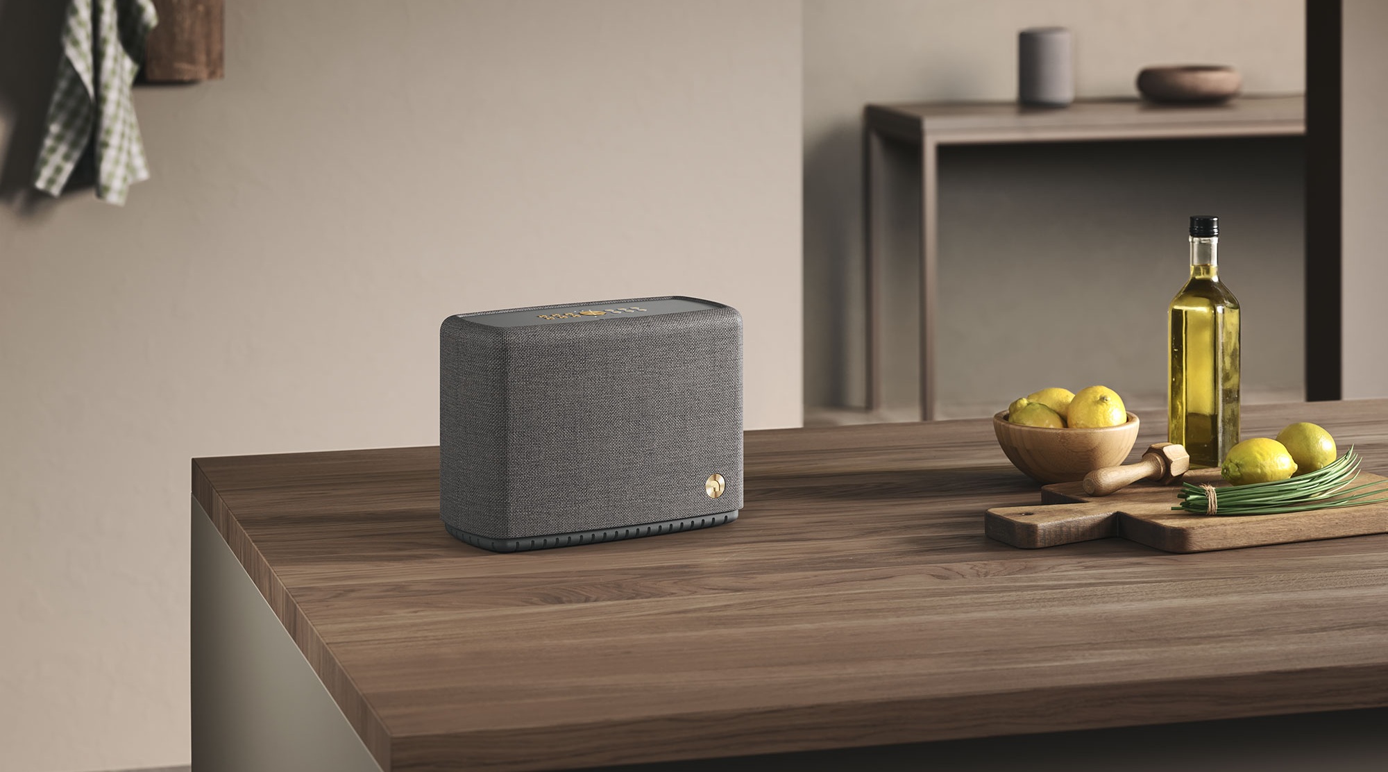 Gray fabric smart speaker sits on a wooden kitchen island beside a bowl of lemons, olive oil bottle, and cutting board.